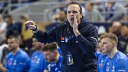 Obrazek przedstawia intensywny moment podczas meczu piłki ręcznej. Mężczyzna w niebieskiej koszulce i granatowych spodenkach, z piłką w prawej ręce, wykonuje rzut lub podanie, będąc w powietrzu. Jest on otoczony przez dwóch przeciwników w jaskrawo-pomarańczowych koszulkach z granatowymi elementami. Zawodnik po jego lewej stronie próbuje blokować jego ruch ręką. Zawodnik po prawej stronie również wyciąga ręce w jego kierunku. W tle widać niewyraźne sylwetki kibiców i elementy hali sportowej (fot. PAP).