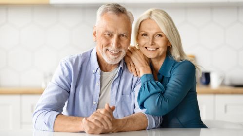 Kobieta i mężczyzna w dojrzałym wieku siedzą przy stole. On opiera dłonie o blat, a ona opiera mu się na ramieniu. Oboje uśmiechają się promiennie.