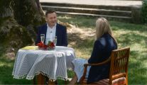 Kadr z programu TVP „Rolnik szuka żony”. Uczestnik Sebastian, ubrany w elegancki garnitur, rozmawia przy stoliku z kobietą o jasnych, długich włosach. Obydwoje znajdują się wśród drzew, zieleni. W tle widać zabytkowe schody.