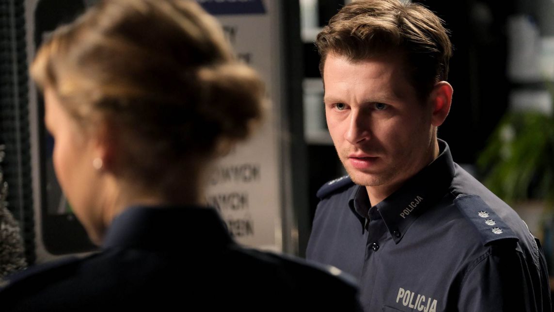 "M jak miłość" - finałowy odcinek sezonu. Co tym razem wydarzy się w serialu? Policjant Adam rozmawia z kobietą widoczną od tyłu. Scena rozgrywa się w ciemnym wnętrzu z tablicą informacyjną w tle.