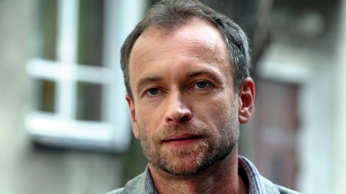 Dr Krzysztof Siennicki, w którego wciela się Mariusz Bonaszewski, w serialu „Glina” patrzy spokojnym, zamyślonym wzrokiem przed siebie. Bohater ma krótko przystrzyżone włosy i lekki zarost, a tło stanowi rozmyty fragment miejskiej zabudowy. Kadr oddaje poważny i refleksyjny nastrój postaci.