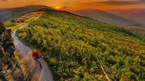 Mężczyzna ubrany w sportowy strój i kask jedzie rowerem po górskim szlaku rowerowym. Krajobraz jest malowniczy. W tle widać górskie szczyty i zachodzące słońce.